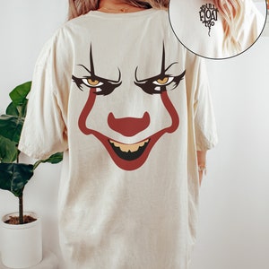 Könnte beinhalten: Elfenbeinfarbenes T-Shirt mit einem großen Clown-Gesicht-Motiv auf dem Rücken, mit roten, schwarzen und gelben Details. Der Text "YOU'LL FLOAT TOO" ist auf dem oberen Rücken aufgedruckt. Das Shirt hat eine lockere Passform.