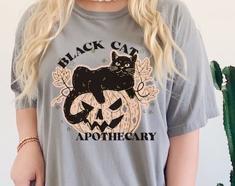 Könnte beinhalten: Ein graues T-Shirt mit einer schwarzen Katze, die auf einem Kürbis sitzt. Der Text "BLACK CAT APOTHECARY" ist auf dem T-Shirt aufgedruckt.