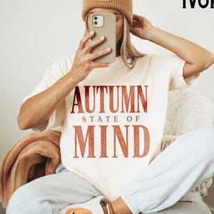 May include: An ivory short-sleeve t-shirt featuring the text AUTUMN STATE OF MIND in a distressed rust-orange font. The shirt is paired with light grey sweatpants and a tan beanie. A white slipper with an orange pumpkin graphic is visible.
