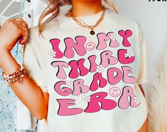 Op de afbeelding: Een licht ivoorwit T-shirt met de tekst "In My Third Grade Era" in roze en witte letters met smiley's en bloemen.