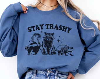 Peut inclure: Un sweat-shirt bleu avec le texte "STAY TRASHY" en lettres noires. Le sweat-shirt présente une illustration en noir et blanc d'un raton laveur, d'une moufette et d'un opossum portant des lunettes de soleil. Le texte "INDIGO BLUE" est dans le coin inférieur droit de l'image.