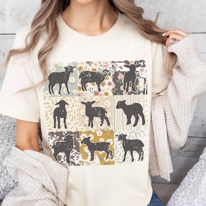 Pode incluir: Uma camiseta de cor creme com um design de patchwork que apresenta nove silhuetas pretas de cordeiros. Cada quadrado do design de patchwork tem um padrão floral diferente em tons de amarelo, verde e azul.