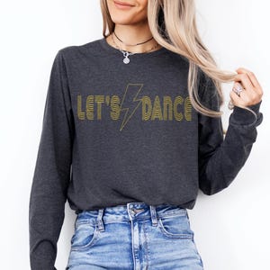Peut inclure: T-shirt à manches longues gris foncé avec l'inscription "LET'S DANCE" en jaune, dans une police rétro, et un éclair jaune. Le t-shirt est porté avec un jean bleu. Les mots "DARK HEATHER" sont dans un encadré blanc en bas à droite.