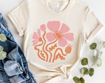 May include: A beige t-shirt with a pink and orange floral design. The design features a large pink flower with a yellow center and smaller pink flowers with orange stems.