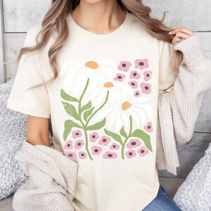 K&ouml;nnte beinhalten: Ein cremefarbenes T-Shirt mit einem grafischen Druck von wei&szlig;en G&auml;nsebl&uuml;mchen und rosa Blumen. Der Text "NATURAL" ist in der oberen rechten Ecke des Bildes sichtbar.
