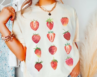 Puede incluir: Una camiseta blanca con un diseño de acuarela de fresas. La camiseta tiene un corte holgado y cuello redondo.