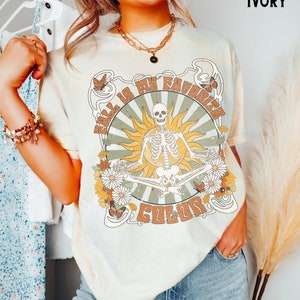 Könnte beinhalten: Ein T-Shirt in Elfenbein mit einem Grafikdruck eines Skeletts, das im Lotussitz mit einer Sonne und Blumen um sich herum sitzt. Der Text "Fall is my favorite color" ist auf dem T-Shirt aufgedruckt.