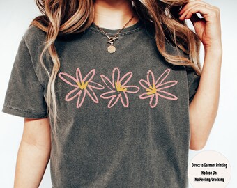 May include: A grey t-shirt featuring a graphic of three pink flowers with yellow centres. The text "Direct to Garment Printing", "No Iron On", and "No Peeling/Cracking" is printed on the bottom right corner of the shirt. The text "PEPPER" is printed in large letters below the text.