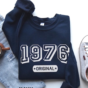 May include: A navy blue sweatshirt featuring the year "1976" in white block lettering, with "ORIGINAL" below. The sweatshirt is displayed on blue denim jeans. A brown mug and a grey trainer are also visible.
