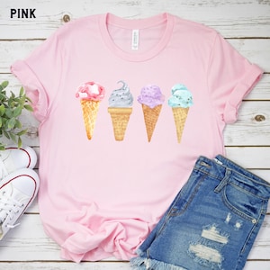 May include: Pink t-shirt with a graphic of four ice cream cones in a row. The ice cream cones are in different colors and flavors, including pink, black, purple, and blue.