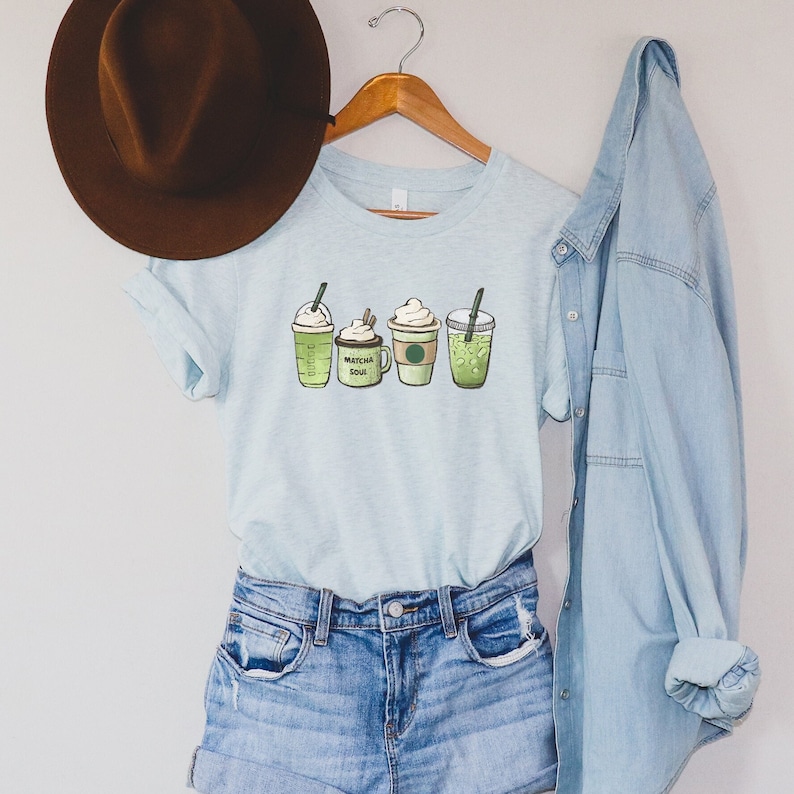 May include: A light blue t-shirt with a graphic of four different green matcha drinks. The drinks are labeled "Matcha Soul" and have whipped cream on top. The shirt is hanging on a wooden hanger with a brown hat and a blue denim jacket.