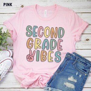 May include: Pink t-shirt with the text "Second Grade Vibes" in colorful letters with white polka dots. A peace sign is in front of the word "Vibes".