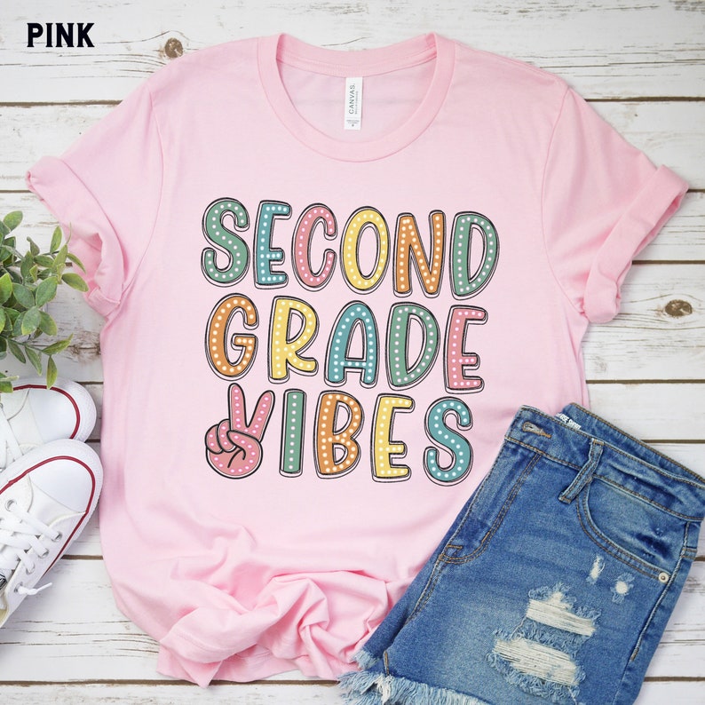 May include: Pink t-shirt with the text "Second Grade Vibes" in colorful letters with white polka dots. A peace sign is in front of the word "Vibes".