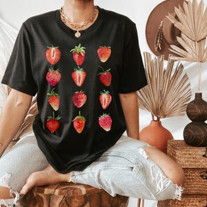 Op de afbeelding: Een zwart T-shirt met een grafische print van aardbeien. De aardbeien zijn rood en hebben groene bladeren. Het shirt wordt gedragen door een persoon.