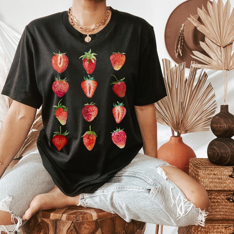 Op de afbeelding: Een zwart T-shirt met een grafische print van aardbeien. De aardbeien zijn rood en hebben groene bladeren. Het shirt wordt gedragen door een persoon.