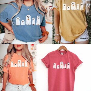May include: Five white ghost dogs wearing Halloween costumes with a pumpkin bucket. The ghosts are printed on a t-shirt in four different colors: blue jean, mustard, terracotta, and crimson.