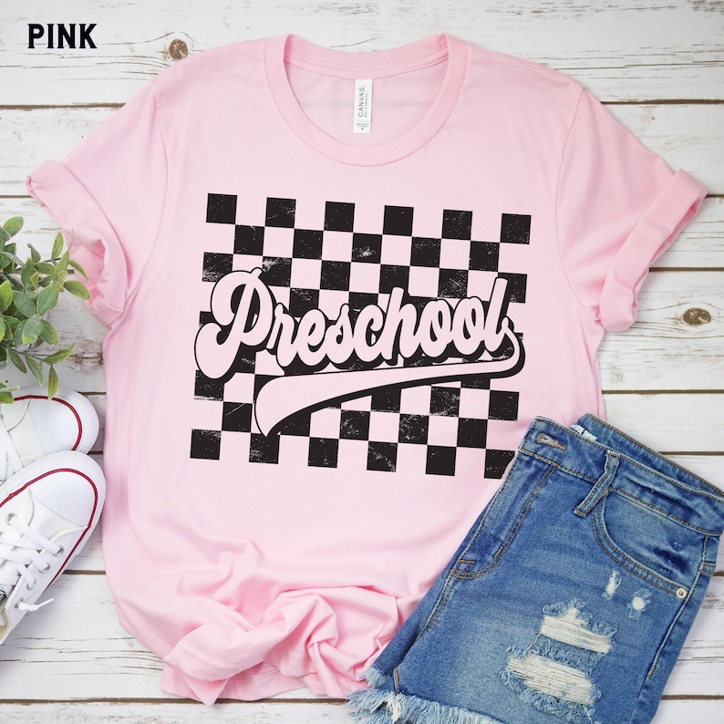 Pu&ograve; includere: T-shirt rosa chiaro con la scritta "Preschool" in un font retr&ograve; su un motivo a scacchi bianco e nero. La maglietta &egrave; abbinata a pantaloncini di jeans e scarpe da ginnastica bianche.