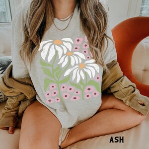 K&ouml;nnte beinhalten: Ein graues Langarmshirt mit einem grafischen Druck von wei&szlig;en G&auml;nsebl&uuml;mchen und rosa Blumen. Das Shirt hat eine lockere Passform und ist aus einem weichen, bequemen Stoff gefertigt.