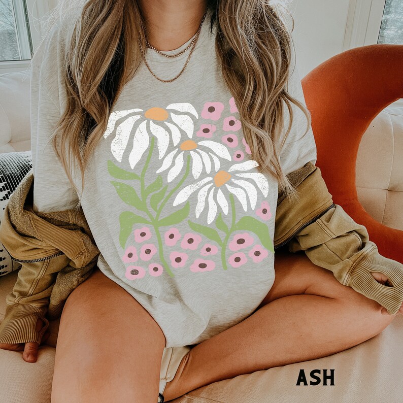 K&ouml;nnte beinhalten: Ein graues Langarmshirt mit einem grafischen Druck von wei&szlig;en G&auml;nsebl&uuml;mchen und rosa Blumen. Das Shirt hat eine lockere Passform und ist aus einem weichen, bequemen Stoff gefertigt.