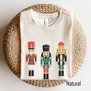 Puede incluir: Una camiseta blanca con un gráfico de tres soldados cascanueces. Los cascanueces llevan uniformes rojos, verdes y azules con detalles dorados. El texto "Natural" está impreso en la esquina inferior derecha de la camiseta.