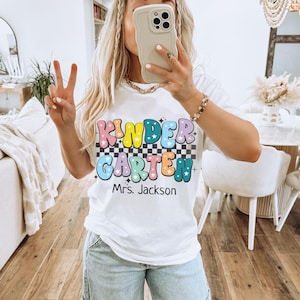 May include: White t-shirt with the text "KINDERGARTEN Mrs. Jackson" in colorful, bubble letters and a checkered background. The shirt is a casual style, suitable for teachers or school staff.