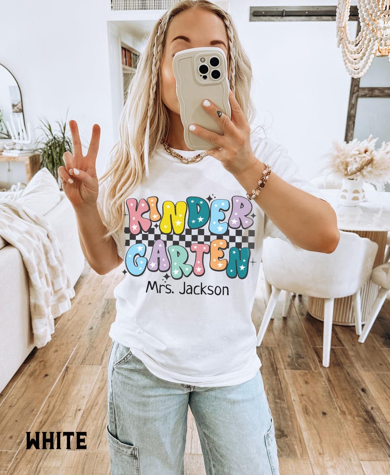 May include: White t-shirt with the text "KINDERGARTEN Mrs. Jackson" in colorful, bubble letters and a checkered background. The shirt is a casual style, suitable for teachers or school staff.