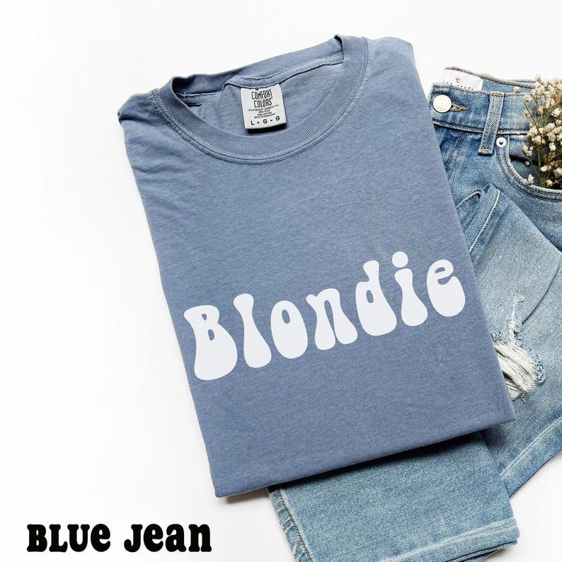 Puede incluir: Una camiseta azul con la palabra "Blondie" en letras blancas onduladas. La camiseta est&aacute; doblada y colocada encima de unos vaqueros azules. El texto "BLUE JEAN" est&aacute; en la parte inferior de la imagen.