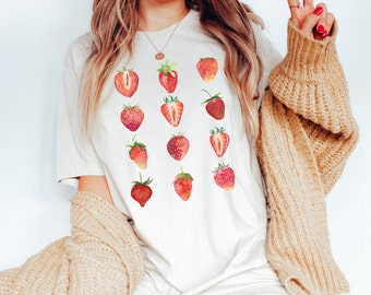 Puede incluir: Una camiseta blanca con un patrón de fresas rojas. Las fresas están dispuestas en una cuadrícula y son de diferentes tamaños y formas.