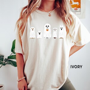 May include: A white t-shirt with a graphic of five ghosts, each with a different dog breed inside. The ghosts are holding a pumpkin bucket and are lined up in a row. The text "IVORY" is in the bottom right corner of the image.