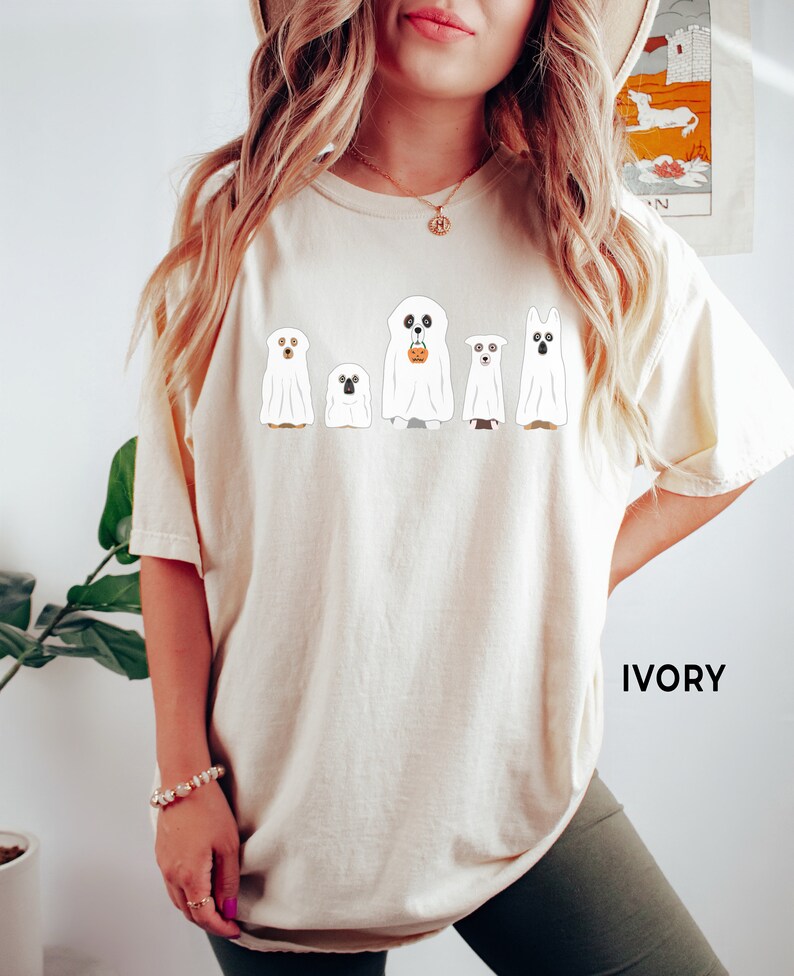 May include: A white t-shirt with a graphic of five ghosts, each with a different dog breed inside. The ghosts are holding a pumpkin bucket and are lined up in a row. The text "IVORY" is in the bottom right corner of the image.