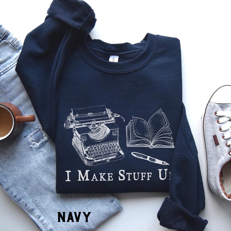Puede incluir: Sudadera azul marino con un gr&aacute;fico blanco de una m&aacute;quina de escribir, un libro abierto y un bol&iacute;grafo. El texto "I MAKE STUFF UP" est&aacute; debajo. La palabra "NAVY" est&aacute; en la parte inferior. La sudadera est&aacute; sobre unos vaqueros azules.