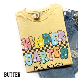 May include: Butter-colored t-shirt with the words "KINDER GARDEN" in colorful bubble letters, with stars and a black and white checkered pattern. The name "Mrs. Jackson" is printed below the words. The shirt is folded on top of blue denim shorts.