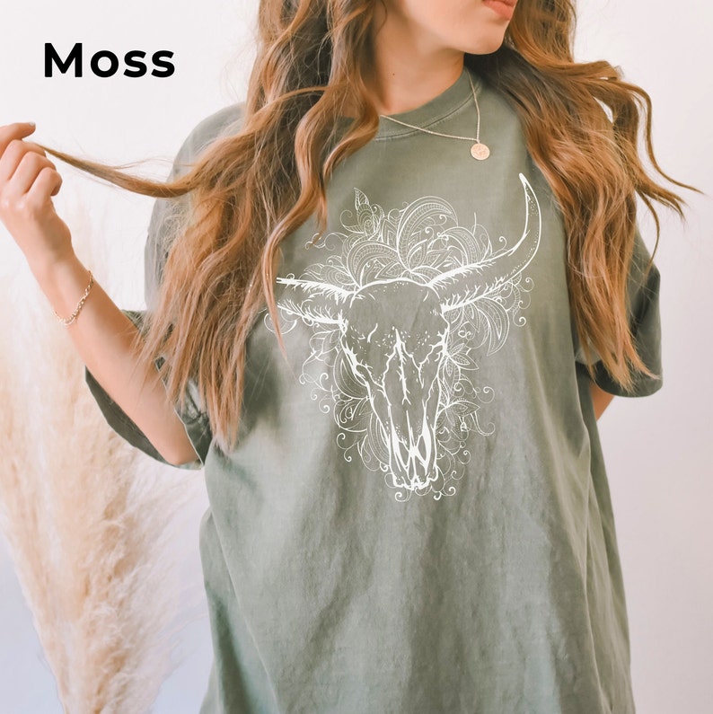K&ouml;nnte beinhalten: Ein gr&uuml;nes Oversize-T-Shirt mit einer wei&szlig;en Grafik eines Bullensch&auml;dels mit floralen Details. Der Text "Moss" befindet sich in der oberen linken Ecke des Bildes.