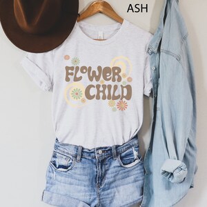 Puede incluir: Una camiseta gris claro con el texto "FLOWER CHILD" en marr&oacute;n, con dise&ntilde;os florales y circulares. La camiseta est&aacute; colgada en una percha de madera, combinada con pantalones cortos de mezclilla y una camisa de mezclilla. Un sombrero marr&oacute;n es visible a la izquierda.