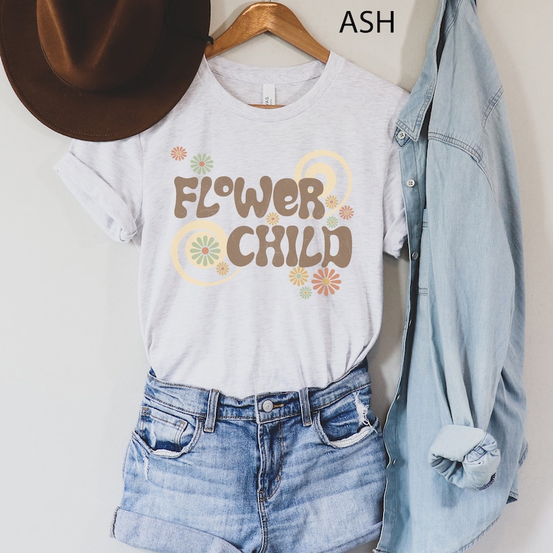 Puede incluir: Una camiseta gris claro con el texto "FLOWER CHILD" en marr&oacute;n, con dise&ntilde;os florales y circulares. La camiseta est&aacute; colgada en una percha de madera, combinada con pantalones cortos de mezclilla y una camisa de mezclilla. Un sombrero marr&oacute;n es visible a la izquierda.