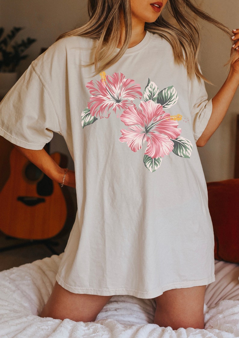 Puede incluir: Una camiseta blanca de gran tama&ntilde;o con estampado floral rosa y verde. Las flores son hibiscos.