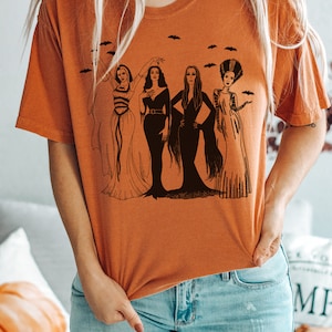 K&ouml;nnte beinhalten: Ein orangefarbenes T-Shirt mit einem Grafikdruck von vier Frauen in schwarzen Kleidern und Frisuren. Die Frauen stehen in einer Reihe und haben unterschiedliche Frisuren und Posen. Die Grafik ist von kleinen schwarzen Flederm&auml;usen umgeben.