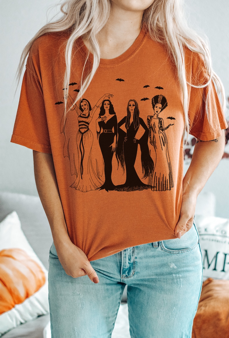 K&ouml;nnte beinhalten: Ein orangefarbenes T-Shirt mit einem Grafikdruck von vier Frauen in schwarzen Kleidern und Frisuren. Die Frauen stehen in einer Reihe und haben unterschiedliche Frisuren und Posen. Die Grafik ist von kleinen schwarzen Flederm&auml;usen umgeben.