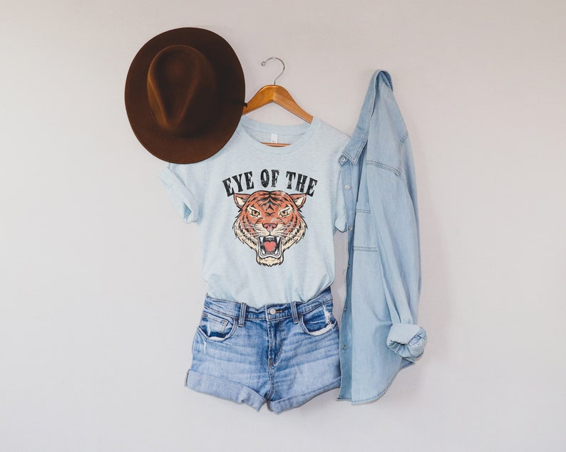 May include: A light blue t-shirt with a tiger graphic and the text "Eye of the Tiger". The shirt is on a hanger with a brown hat and a light blue denim jacket.