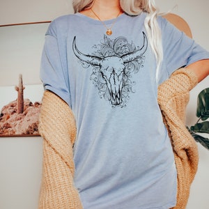 Peut inclure: Un t-shirt bleu clair avec un motif noir et blanc de cr&acirc;ne de taureau avec des accents floraux.