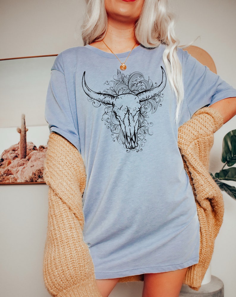 Peut inclure: Un t-shirt bleu clair avec un motif noir et blanc de cr&acirc;ne de taureau avec des accents floraux.