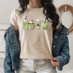 May include: A beige t-shirt with a graphic of five different green drinks. The drinks include a coffee, a matcha latte, a green tea, and two iced coffees. The text "Matcha Soul" is on the graphic.