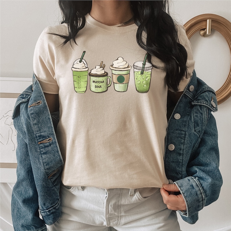 May include: A beige t-shirt with a graphic of five different green drinks. The drinks include a coffee, a matcha latte, a green tea, and two iced coffees. The text "Matcha Soul" is on the graphic.