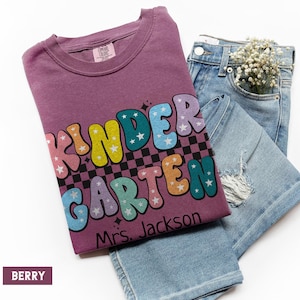 May include: A berry-colored t-shirt with the word "KINDERGARTEN" in colorful, bubble letters, with stars and a checkered pattern. The text "Mrs. Jackson" is below the word. Light blue denim jeans are folded next to the shirt.