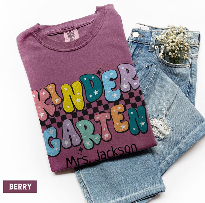 May include: A berry-colored t-shirt with the word "KINDERGARTEN" in colorful, bubble letters, with stars and a checkered pattern. The text "Mrs. Jackson" is below the word. Light blue denim jeans are folded next to the shirt.