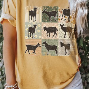 May include: A mustard yellow t-shirt with a patchwork design featuring silhouettes of goats. The design includes various floral patterns and geometric shapes.