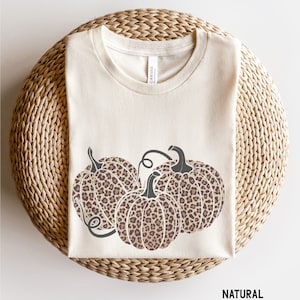May include: A beige t-shirt featuring a graphic of three brown and black leopard print pumpkins. The text "NATURAL" is printed at the bottom of the shirt.