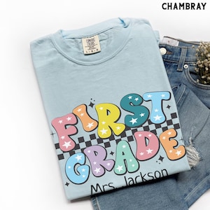 May include: Light blue t-shirt with the words "FIRST GRADE" in colorful bubble letters with star accents. The shirt also includes the name "Mrs. Jackson" in black text. The shirt is folded on top of denim shorts.