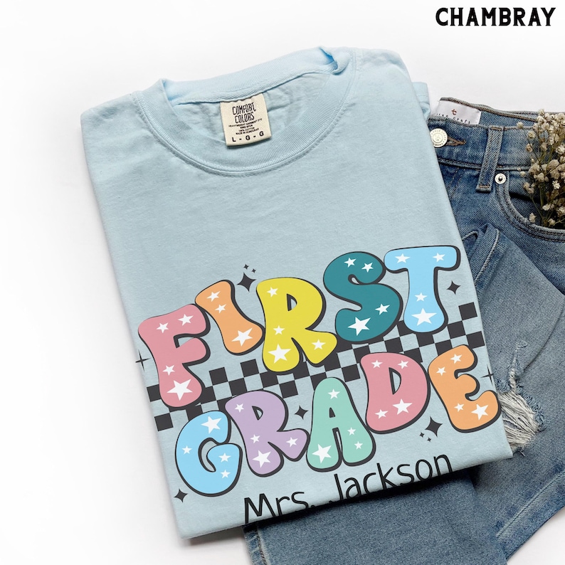 May include: Light blue t-shirt with the words "FIRST GRADE" in colorful bubble letters with star accents. The shirt also includes the name "Mrs. Jackson" in black text. The shirt is folded on top of denim shorts.