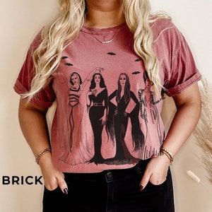 K&ouml;nnte beinhalten: Ein maronfarbenes T-Shirt mit einem grafischen Druck von vier Frauen in schwarzen Kleidern vor wei&szlig;em Hintergrund. Die Frauen tragen schwarze Kleider mit wei&szlig;en Akzenten und haben langes schwarzes Haar. Das Shirt hat einen Rundhalsausschnitt und kurze &Auml;rmel.
